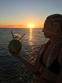 Woman holding umbrella by sea against sky during sunset