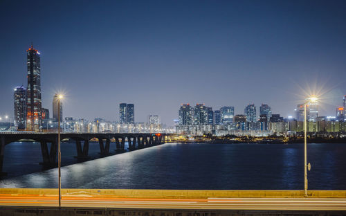 Illuminated bridge over river by buildings against sky at night