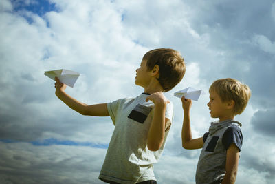 Side view of siblings playing with paper airplanes against cloudy sky