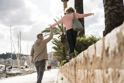 Mature man holding hand of woman walking on wall by harbor