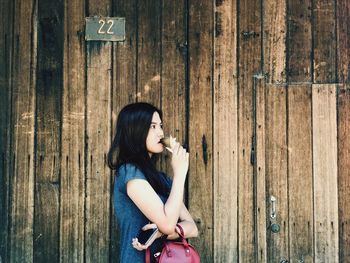 Side view of thoughtful woman eating ice cream cone against wooden wall