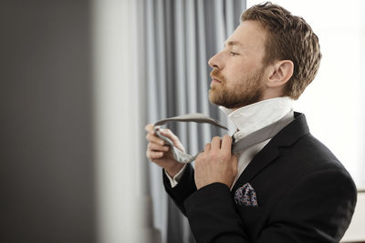 Side view of businessman fastening necktie against window in hotel room