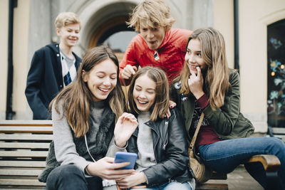 Happy friends looking at smart phone while sitting on bench at city