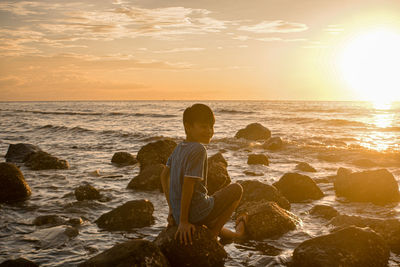 Rear view of man on rock at beach during sunset