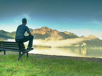 Alone man sits on bench beside an azure mountain lake. man relax and watch high peaks of alps above 