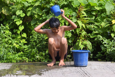 A rural child taking a bath by the bucket on the road, plants and greenery in the background
