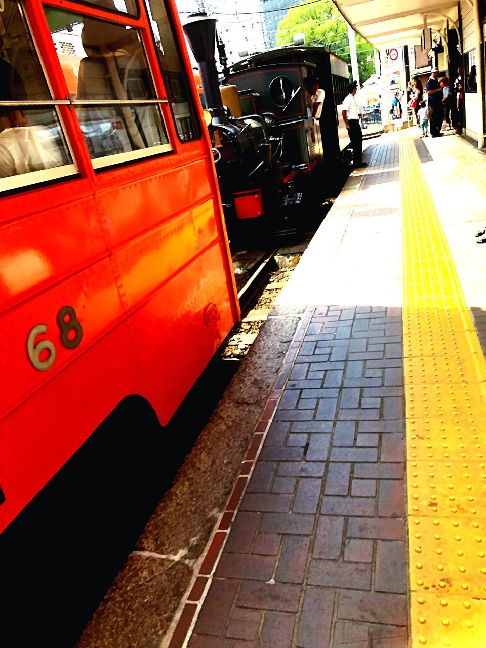 transportation, city, mode of transportation, street, land vehicle, public transportation, incidental people, day, architecture, red, outdoors, footpath, road, travel, motor vehicle, built structure, car, real people, railroad station, city life