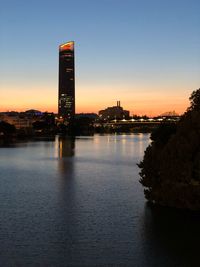 Illuminated buildings by river against sky during sunset