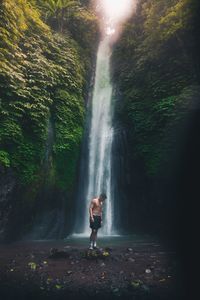 Full length of shirtless man standing against waterfall in forest