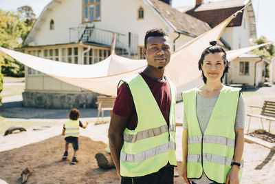 Portrait of confident mid adult male and female teachers standing in reflective clothing at playground