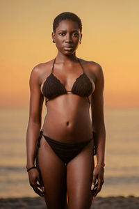 Portrait of young woman in bikini standing at beach during sunset