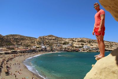 Full length of man standing on cliff by sea against clear blue sky
