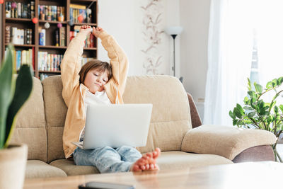 The girl sits on the couch in front of a laptop and stretches from fatigue. children use technology