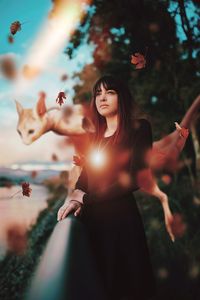 Digital composite image of thoughtful young woman looking away while standing against trees during sunset