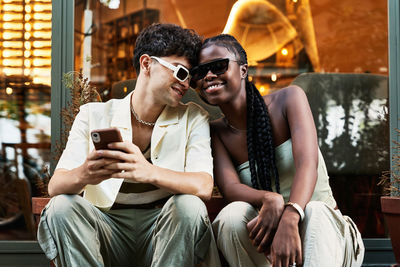 Happy black woman in stylish clothes and sunglasses smiling and leaning on boyfriend using smartphone while sitting against glass wall on city street