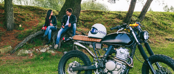 Young couple resting from a motorcycle trip having a beer outdoors with motorbike in foreground