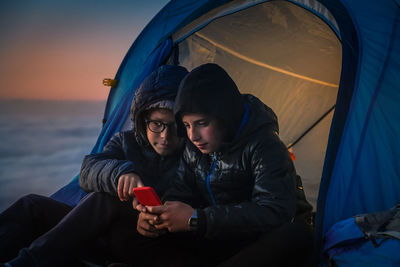 Brothers using mobile phone while sitting in tent at sunset