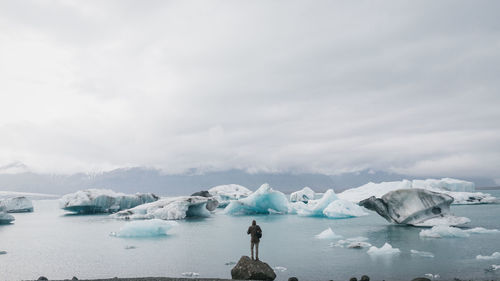 Rear view of man standing on rock against icebergs in sea against sky