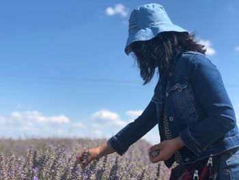 Side view of woman picking lavenders from farm against blue sky