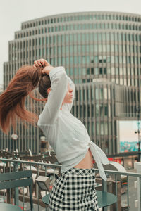 Side view of young woman holding hair while standing by buildings in city