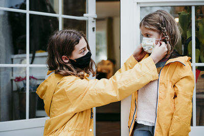 Sister adjusting face mask of sister while sitting against door