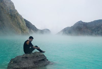 Man sitting on rock in sea during foggy weather