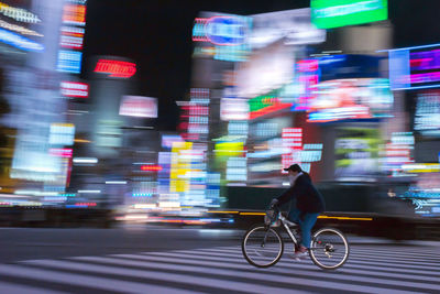 Side view of woman riding bicycle on street in illuminated city at night