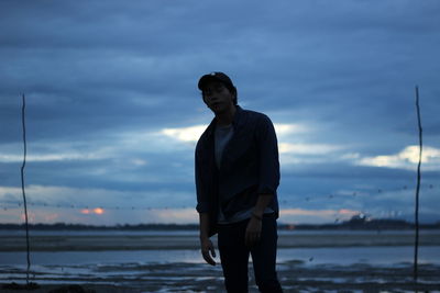 Young man standing at lakeshore against cloudy sky