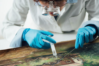 A female conservator with magnifying goggles repairing damages on an oil painting canvas.