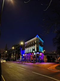 Illuminated street by buildings against sky at night