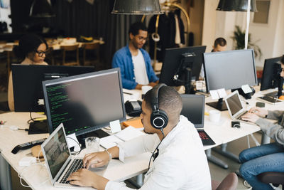 High angle view of businessman with headphones coding over laptop while coworkers working in background