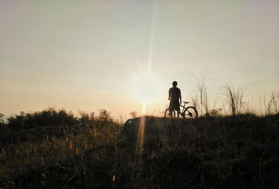 Silhouette young man with bicycle standing on field against sky during sunset