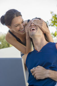 Happy woman covering man eyes in city against sky
