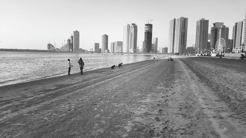 People walking on beach by cityscape against clear sky