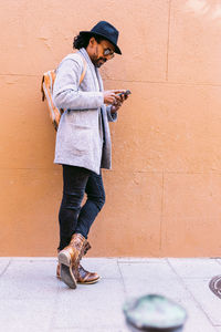 Full body hispanic male in stylish clothes with hat and sunglasses leaning on wall and browsing cellphone while standing on pavement on city street