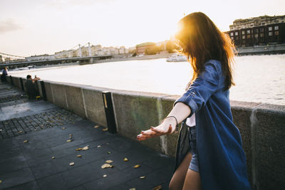 Woman standing on sidewalk by river against sky during sunset