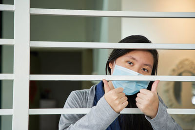 Portrait of young woman with face mask with thums up behind window