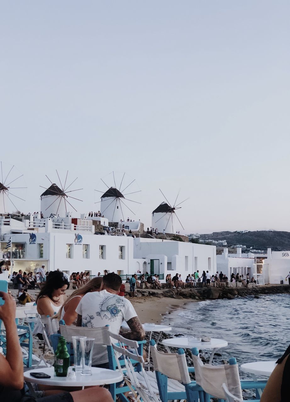 PEOPLE SITTING BY SEA AGAINST CLEAR SKY WITH BUILDINGS IN BACKGROUND