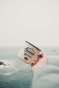 Cropped hand of person holding cup by sea against sky