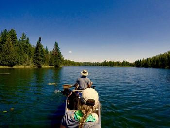 Rear view of friends canoeing with dog on lake against blue sky