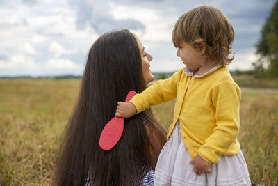 Adorable toddler girl play combing her mom's hair outdoor. rear view of mother and daughter on field