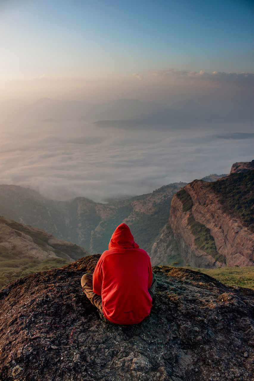 rear view, beauty in nature, sky, scenics - nature, tranquility, tranquil scene, mountain, one person, rock, non-urban scene, real people, rock - object, solid, cloud - sky, nature, red, idyllic, environment, sitting, remote, mountain range, outdoors, hood - clothing, looking at view