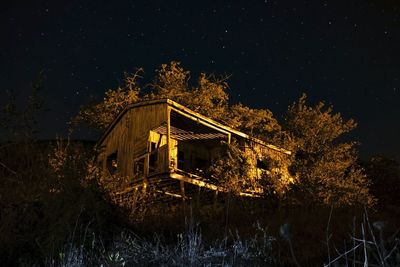 Illuminated house on field by trees against sky at night