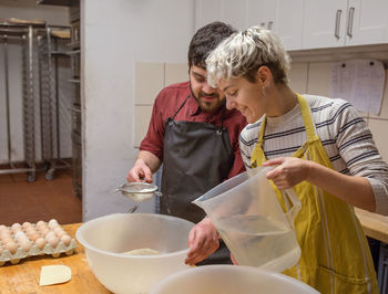 Couple of baker sifting flour and pouring water into bowl while preparing dough for bread during work on kitchen of bakery