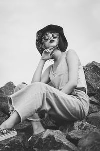 Young woman looking away while sitting on rock against sky