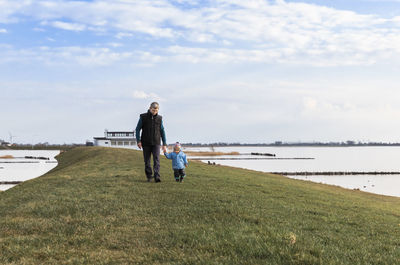 Grandfather and granddaughter walking at lakeshore against sky