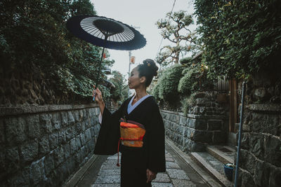 Mature woman holding umbrella standing by plants amidst alley