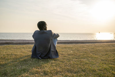 Young businessman sitting on grass and admiring sea at sunset