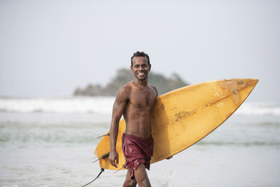 Portrait of shirtless male surfer carrying surfboard in sea