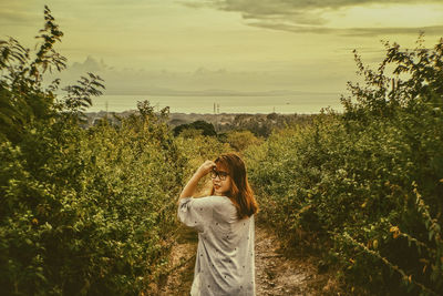 Portrait of young woman standing by plants against sky